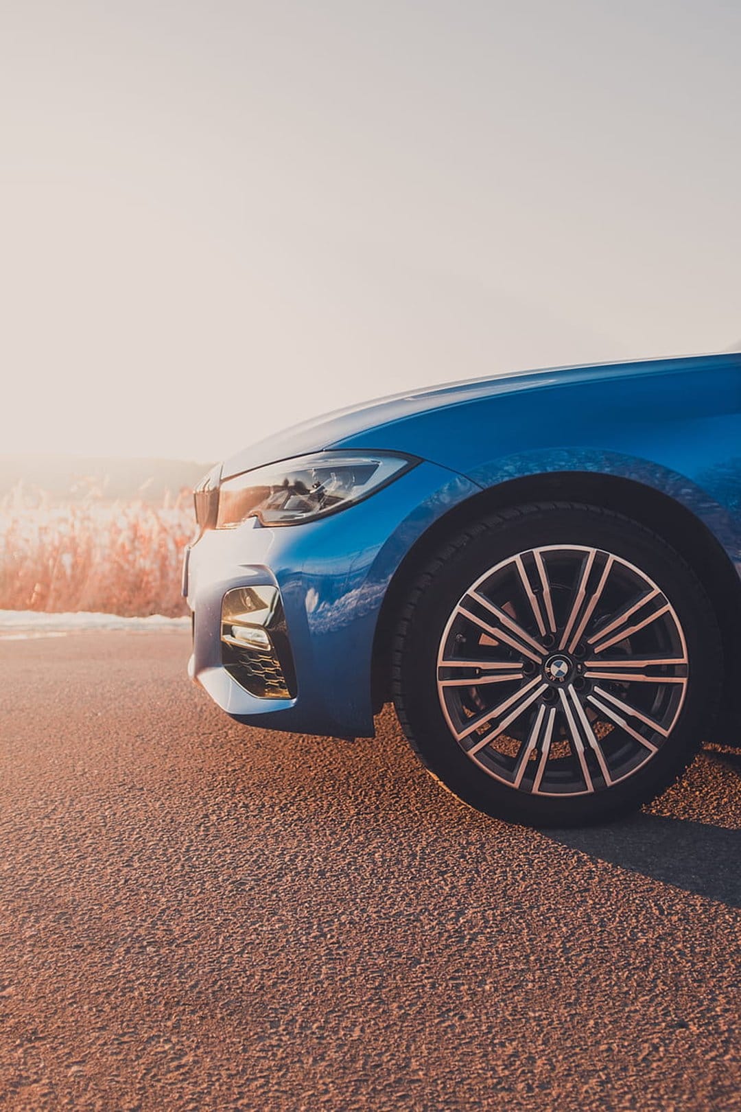 Blue BMW car parked on asphalt with sunset light hitting the front wheel and tall grass in the background.