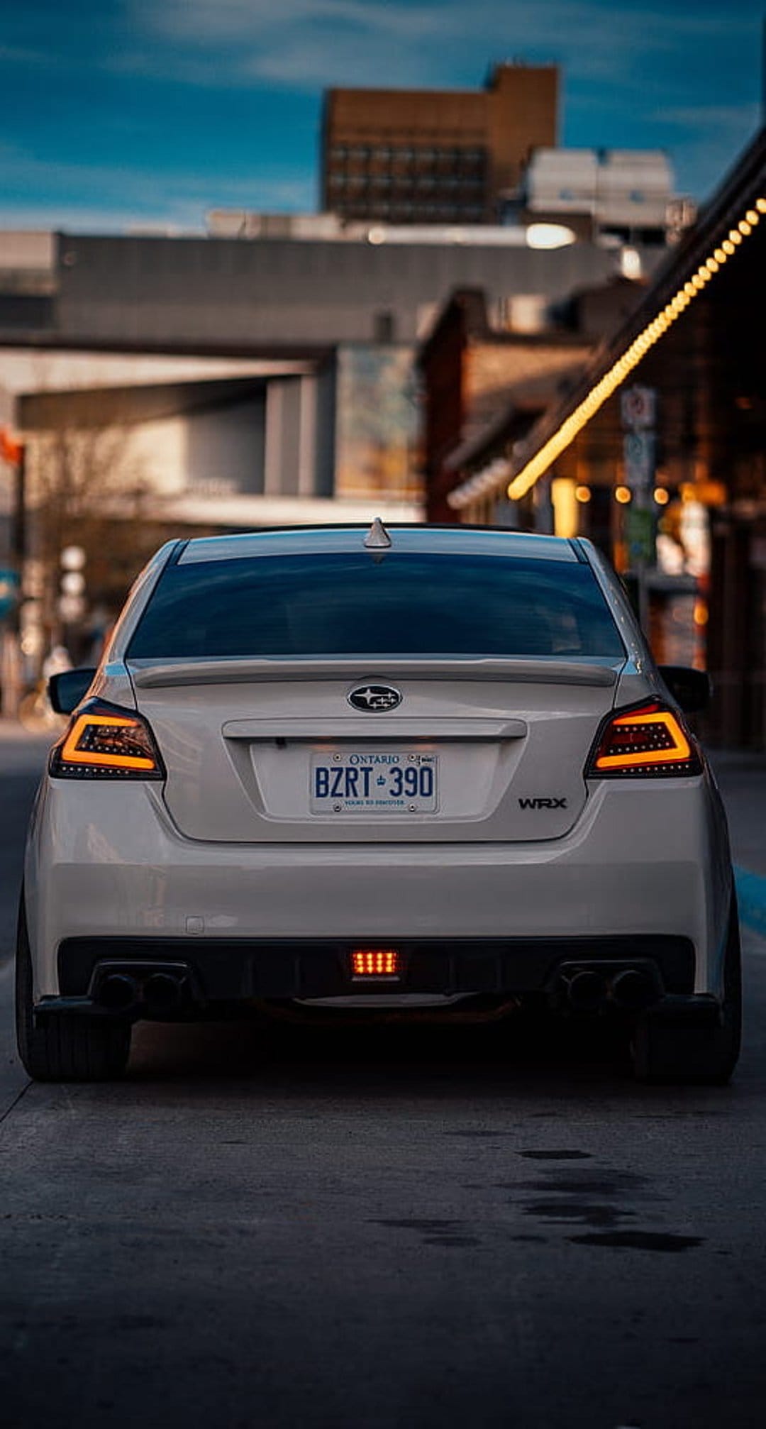Subaru WRX rear view with glowing red LED taillights on a wet asphalt road under urban night city lights.