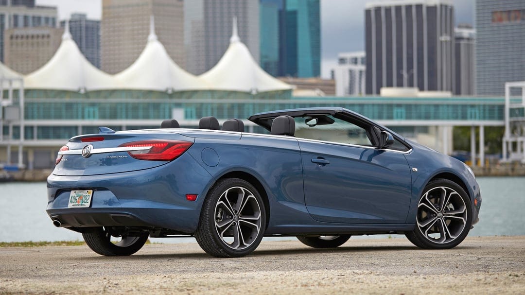 Blue Buick Cascada convertible parked on concrete with an urban skyline and white tents in the background.