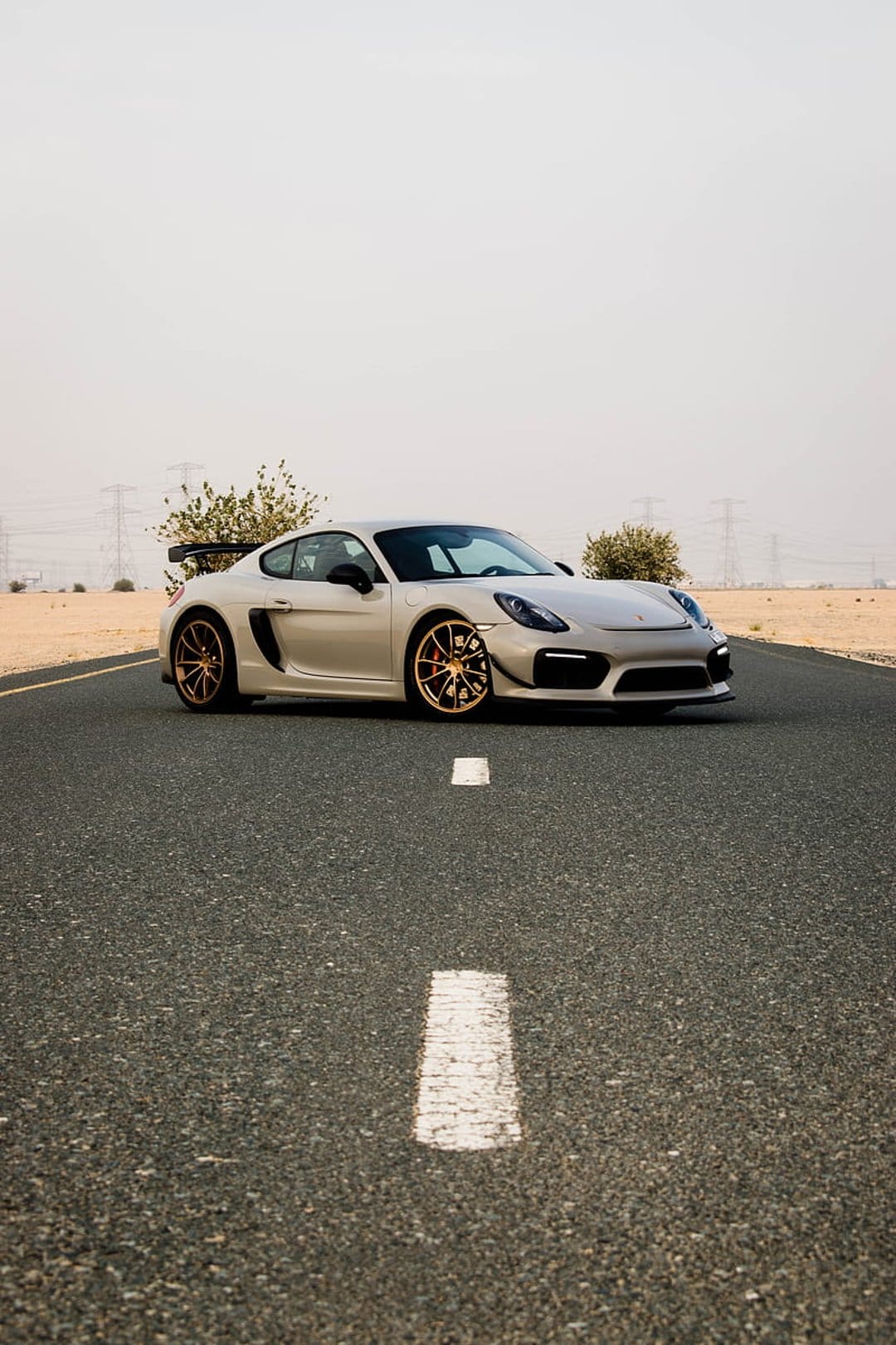 Silver Porsche Cayman GT4 with gold wheels on a desert asphalt road under bright sunlight with a rear spoiler.