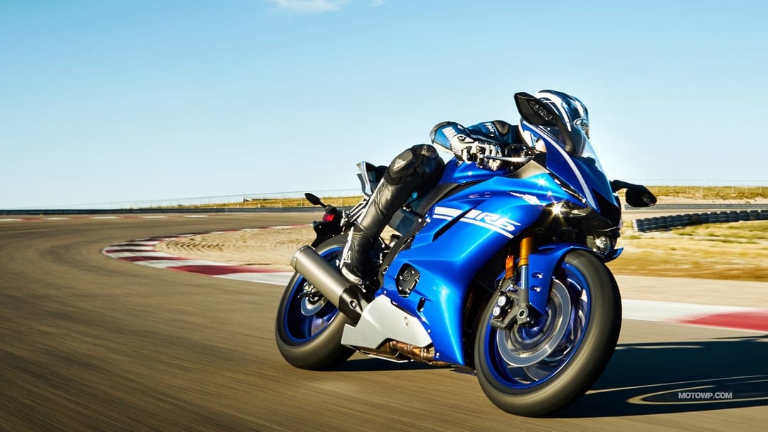 Blue Yamaha YZF-R6 sportbike leaning into a racetrack turn with a rider in full leathers under a clear sky.