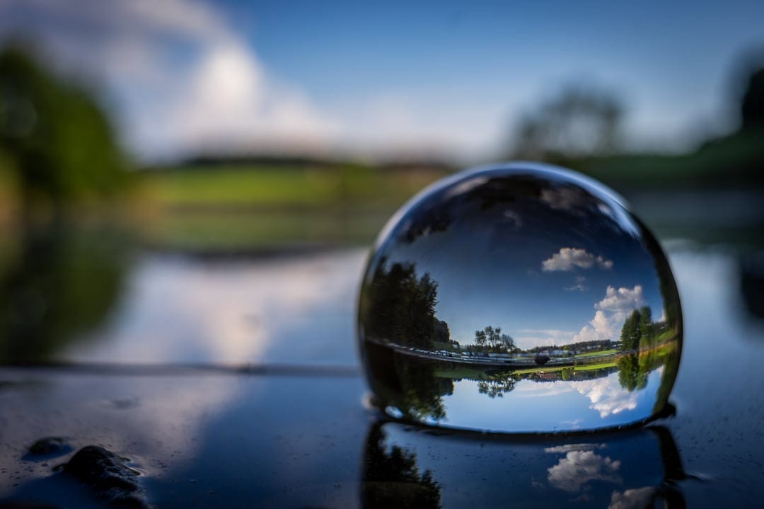 Crystal sphere on a dark wet floor reflects an inverted forest lake under a bright blue cloudy sky.