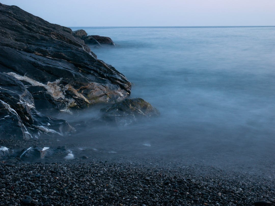Jagged dark rocks and smooth pebbles meet a misty, long-exposure ocean shore under a soft twilight haze.