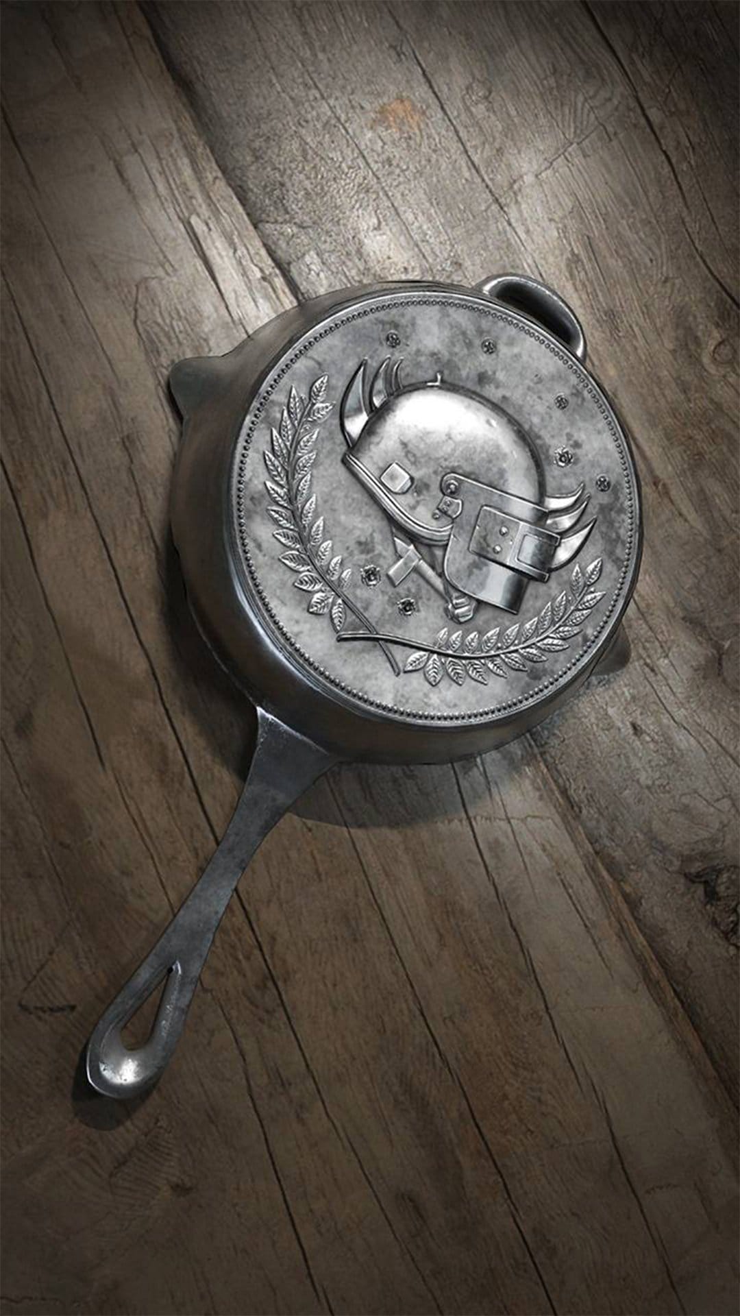 Silver frying pan with a helmet emblem and bullet holes rests on a rustic wooden surface in dramatic lighting.
