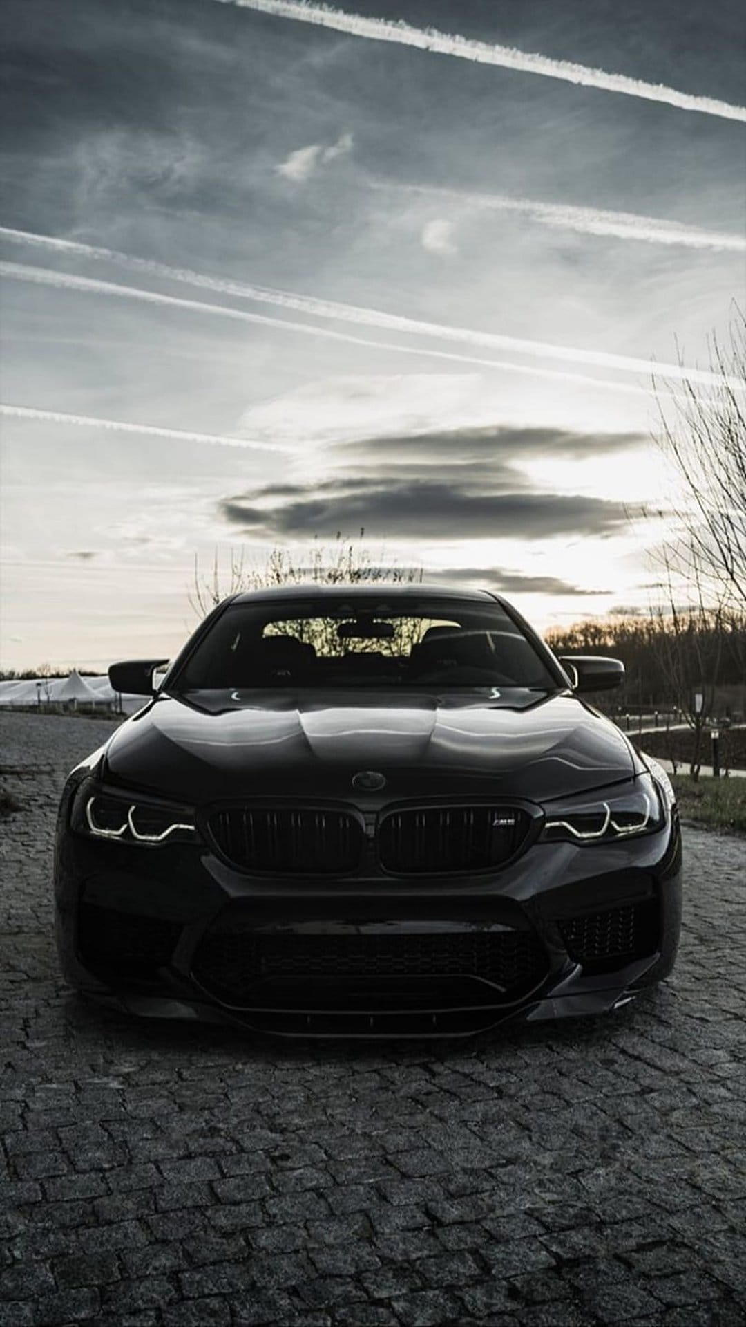 Black BMW M5 parked on a cobblestone road beneath a cloudy sky with white contrails and dark tree branches.