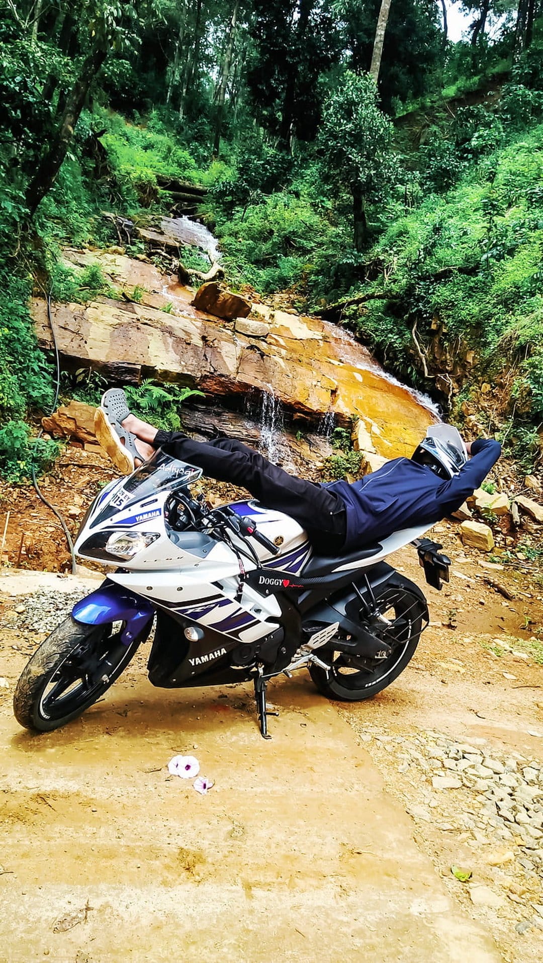 Biker in blue gear rests on a white motorcycle parked by a rocky woodland waterfall amidst dense green ferns.