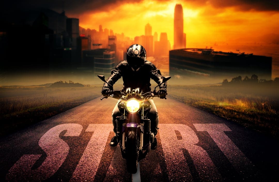Motorcyclist in leather gear riding past a white start sign on a road against a fiery orange sunset skyline.