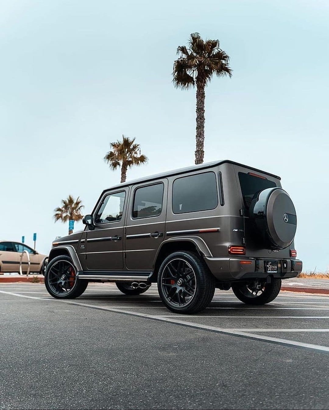 Dark grey G-Wagon SUV parked on asphalt near tropical palm trees and a beach under a clear pale blue sky.