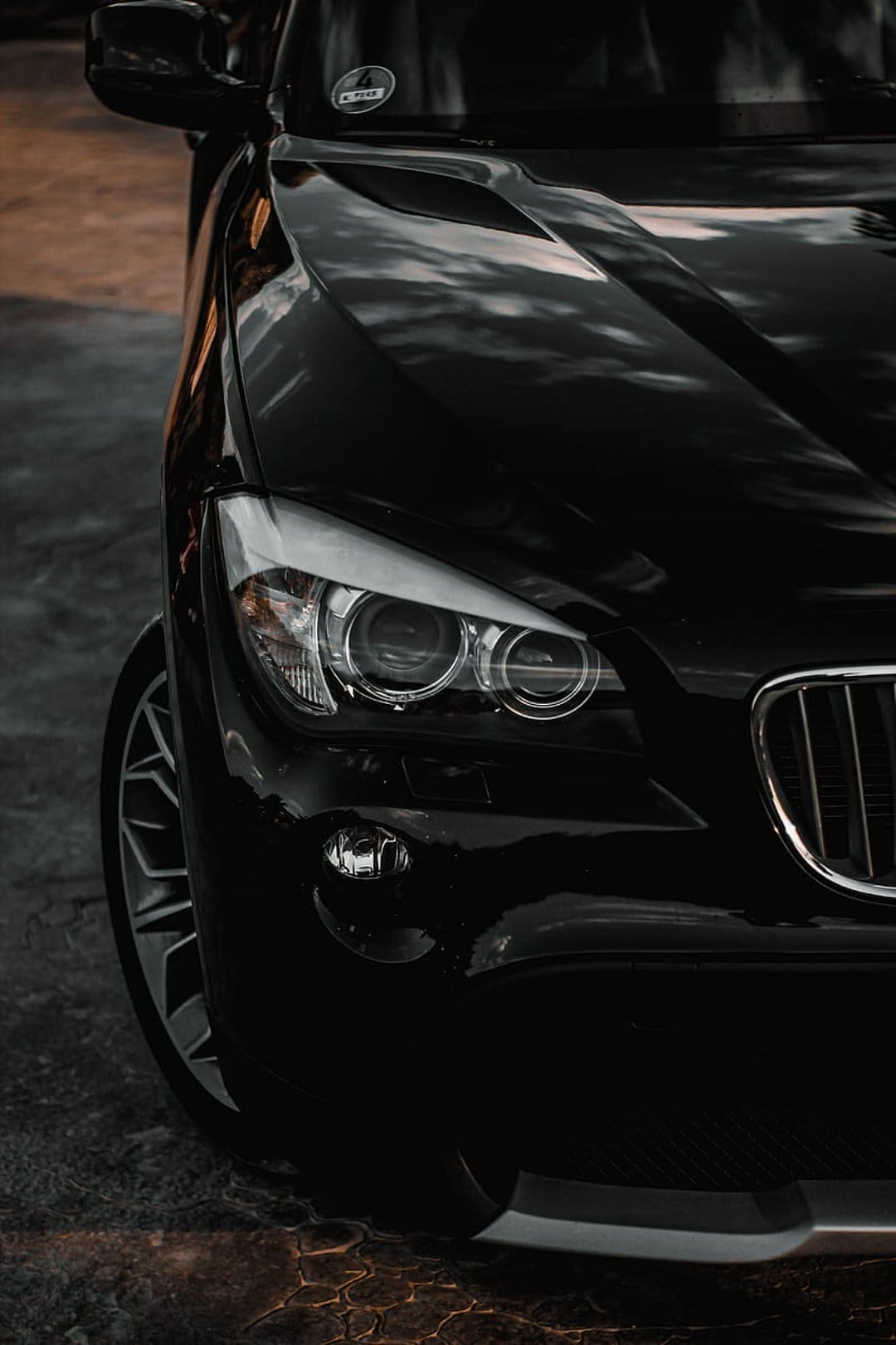 Black BMW X1 headlight and kidney grille with reflections of a cloudy sky above a textured pavement surface.