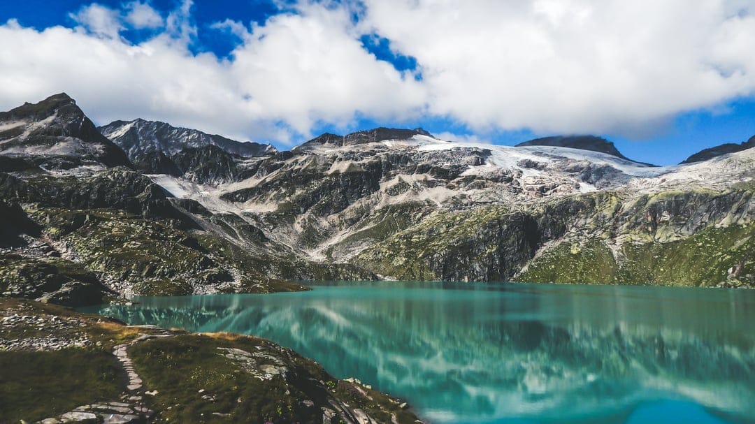 Turquoise alpine lake reflecting snow-capped mountain peaks under a blue sky with soft white cumulus clouds.