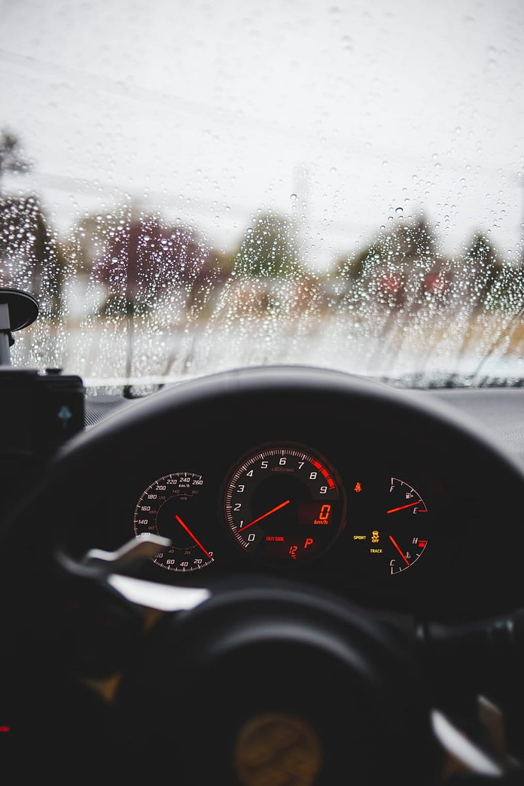 Car dashboard with glowing red gauges viewed through a windshield covered in rain droplets and gray mist.