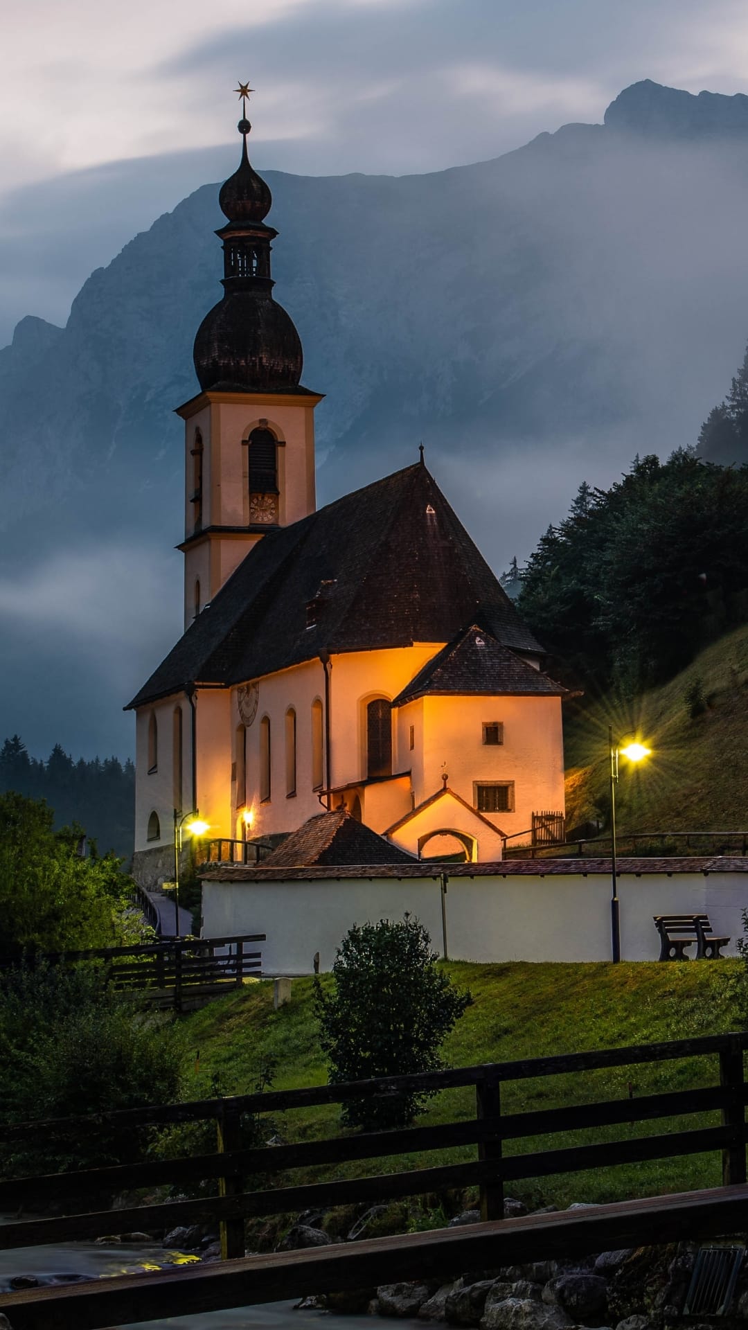 St. Sebastian Church with an illuminated steeple stands before misty mountains beside a flowing creek at dusk.