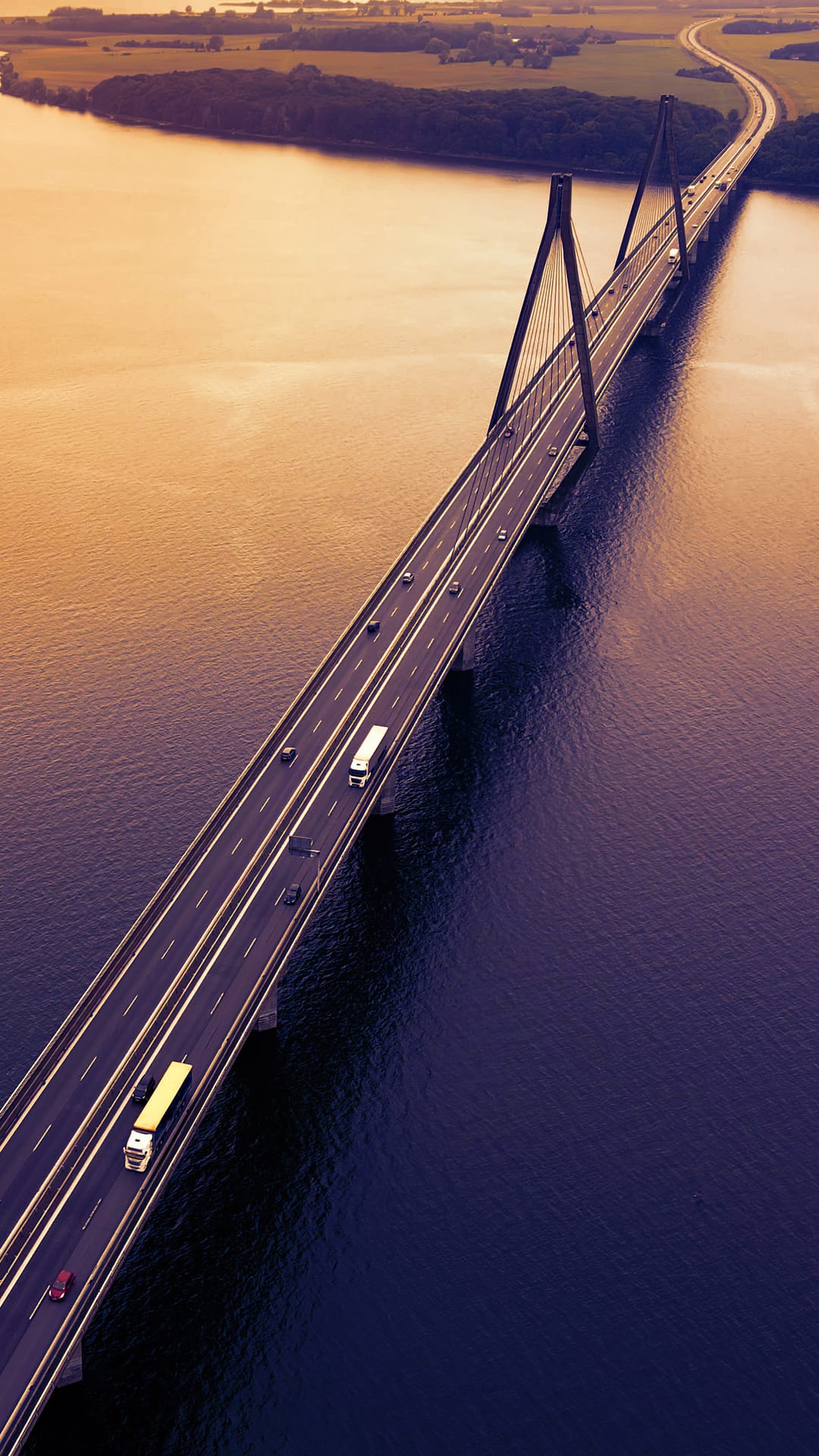 Cable-stayed bridge spanning golden water at sunset with small cars driving along the distant highway lanes.