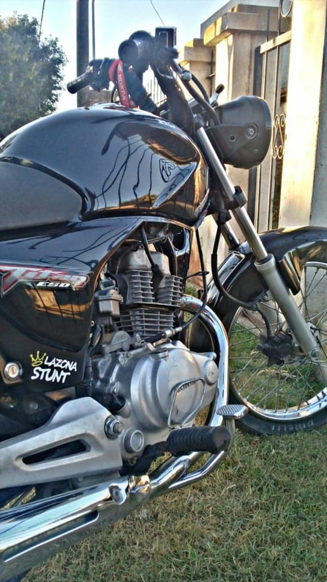 Black Honda motorcycle engine and fuel tank with a Lazona Stunt sticker on a sunny day near green grass.