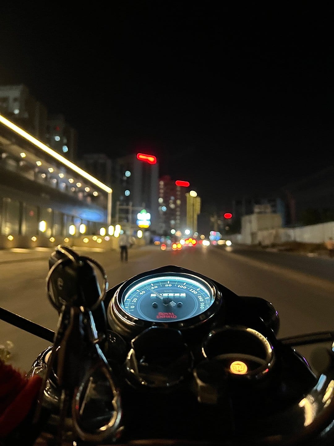 Royal Enfield motorcycle speedometer with glowing orange needles against a blurred nighttime city street backdrop.