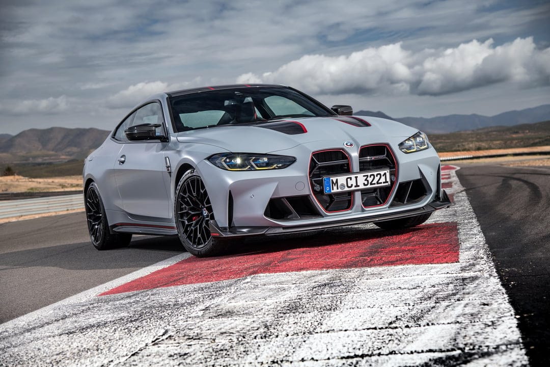 Grey BMW M4 CSL with red grille accents parked on a racetrack curb under a cloudy blue and white sky.