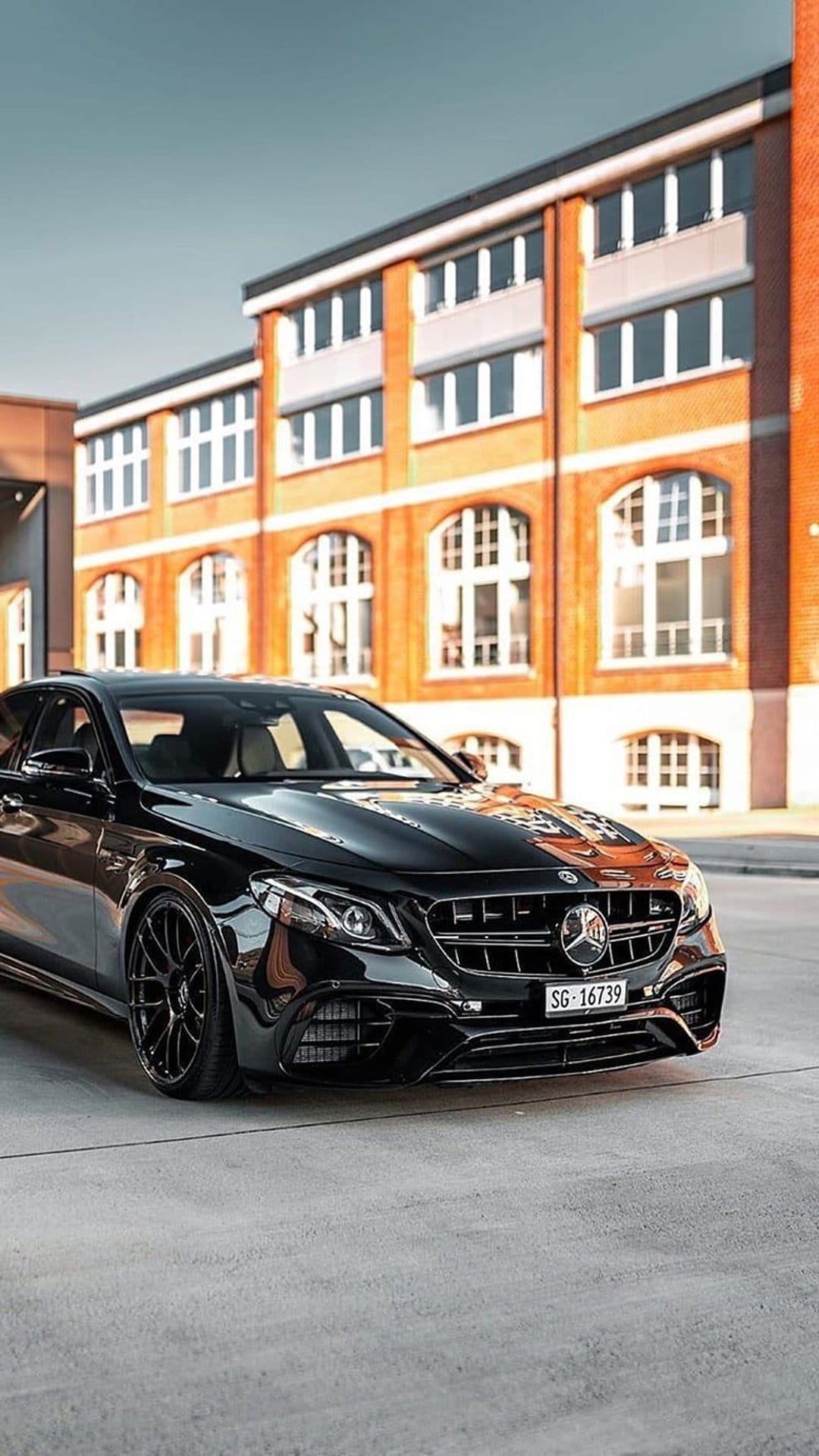 Black Mercedes-AMG E63s sedan parked by a brick building with custom wheels and sharp headlights visible.