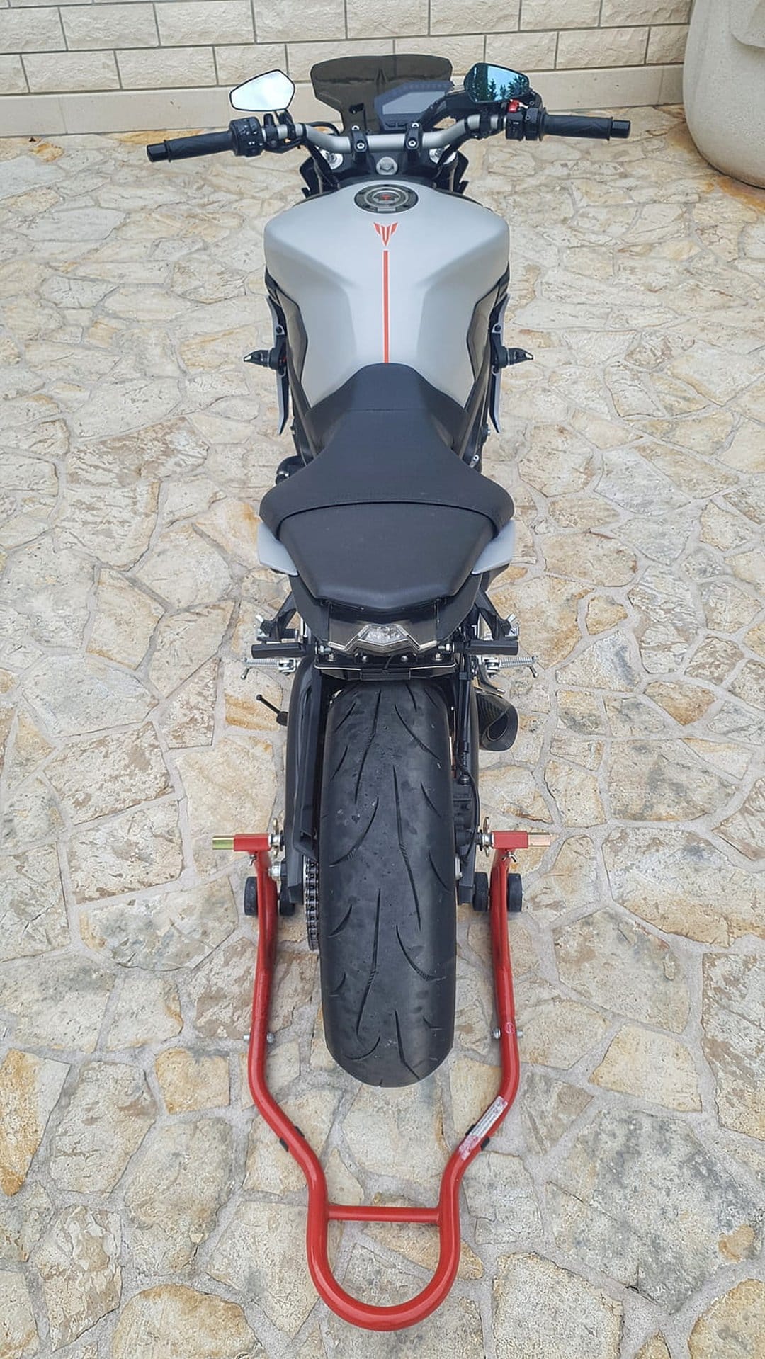 Yamaha MT-09 motorcycle rear view on a red stand with a wide tire, silver bodywork, and a brick wall background.