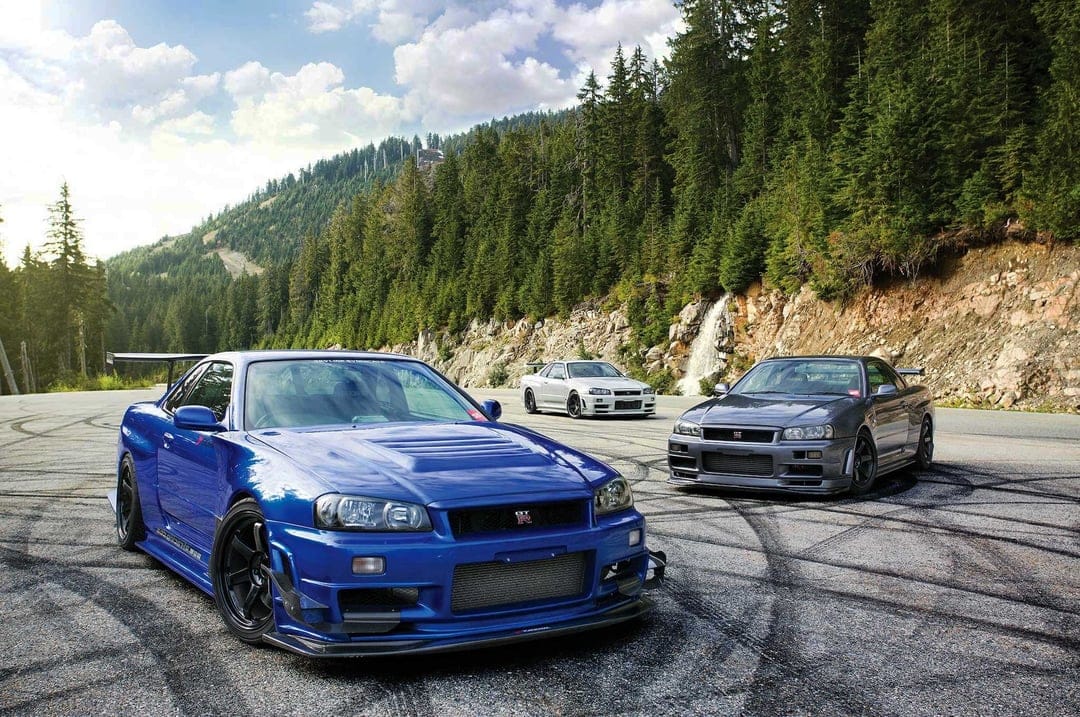 Three Nissan Skyline GT-R R34 cars parked on a mountain road with tire marks against a forest backdrop.