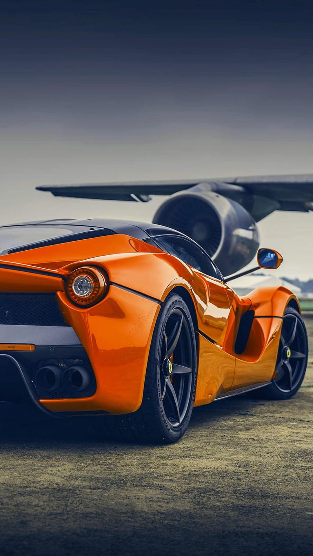 Orange Ferrari parked beside a large silver airplane turbine engine under soft lighting in a portrait composition.