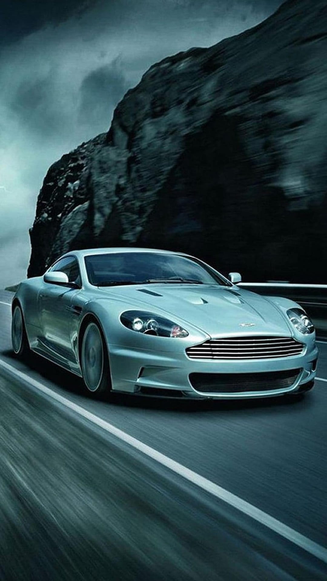 Silver Aston Martin DB9 speeds past a rocky cliff on a winding road under a moody, overcast sky with motion blur.
