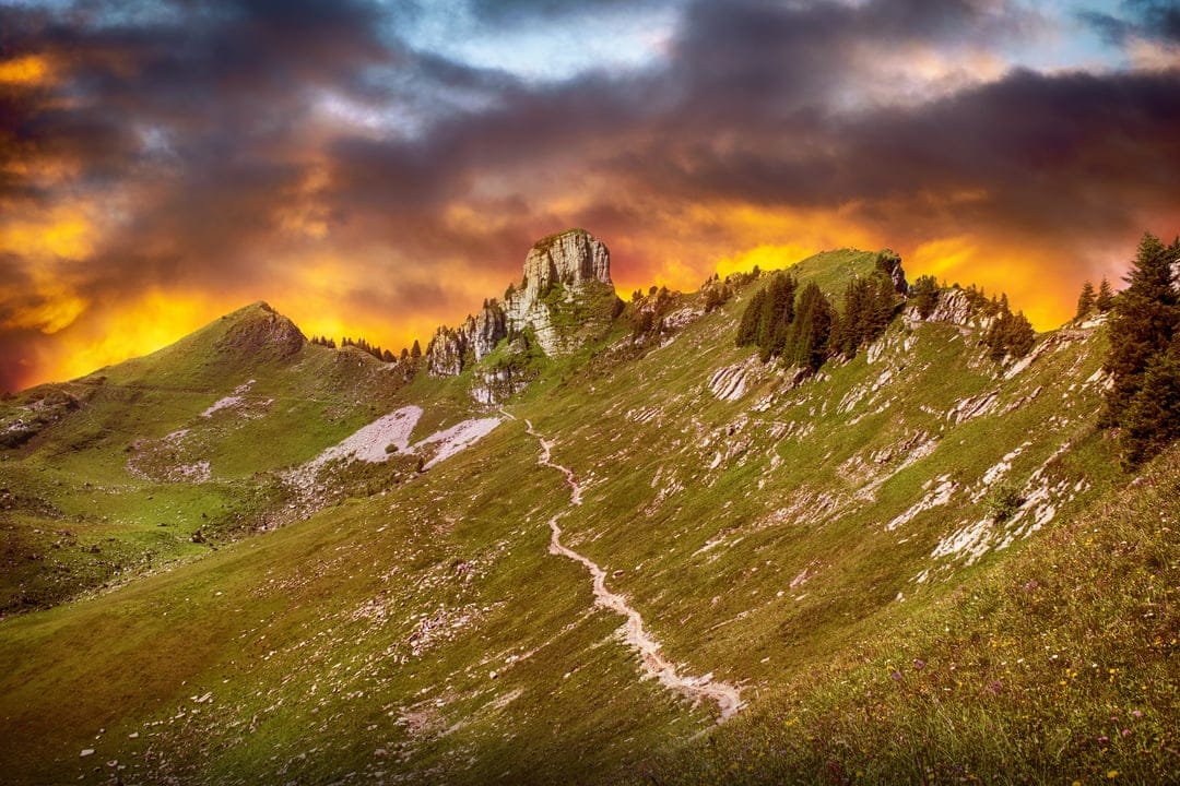 Jagged mountain peak under a purple and orange sky with a narrow hiking path through green grassy slopes.