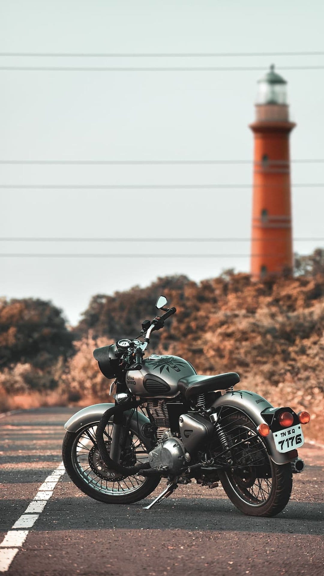 Silver vintage motorcycle parked on a coastal road leading toward a tall orange lighthouse under soft light.