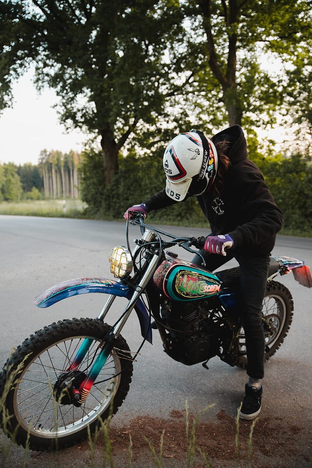 Motocross rider in a white helmet and black hoodie sits on a dirt bike with a colorful mural gas tank.