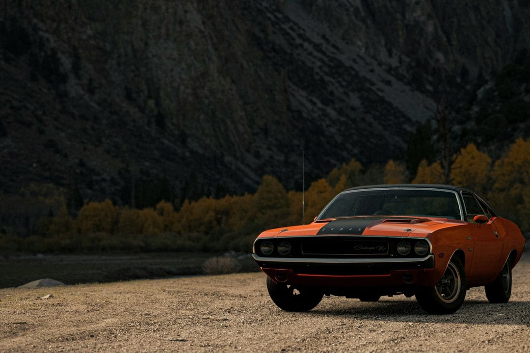 Orange Dodge Challenger parked on a gravel mountain road during autumn with dark, moody peaks in the distance.
