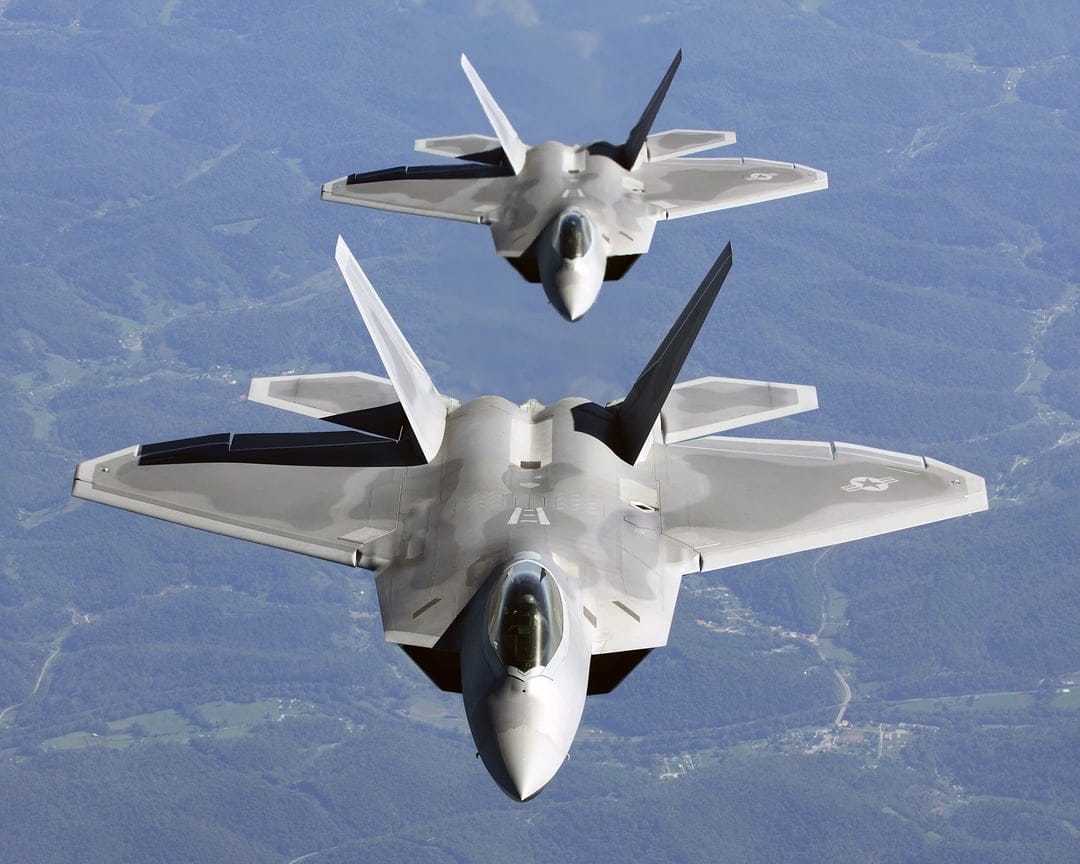 Two F-22 Raptor stealth jets fly in formation over a blurred mountainous landscape with delta wings visible.
