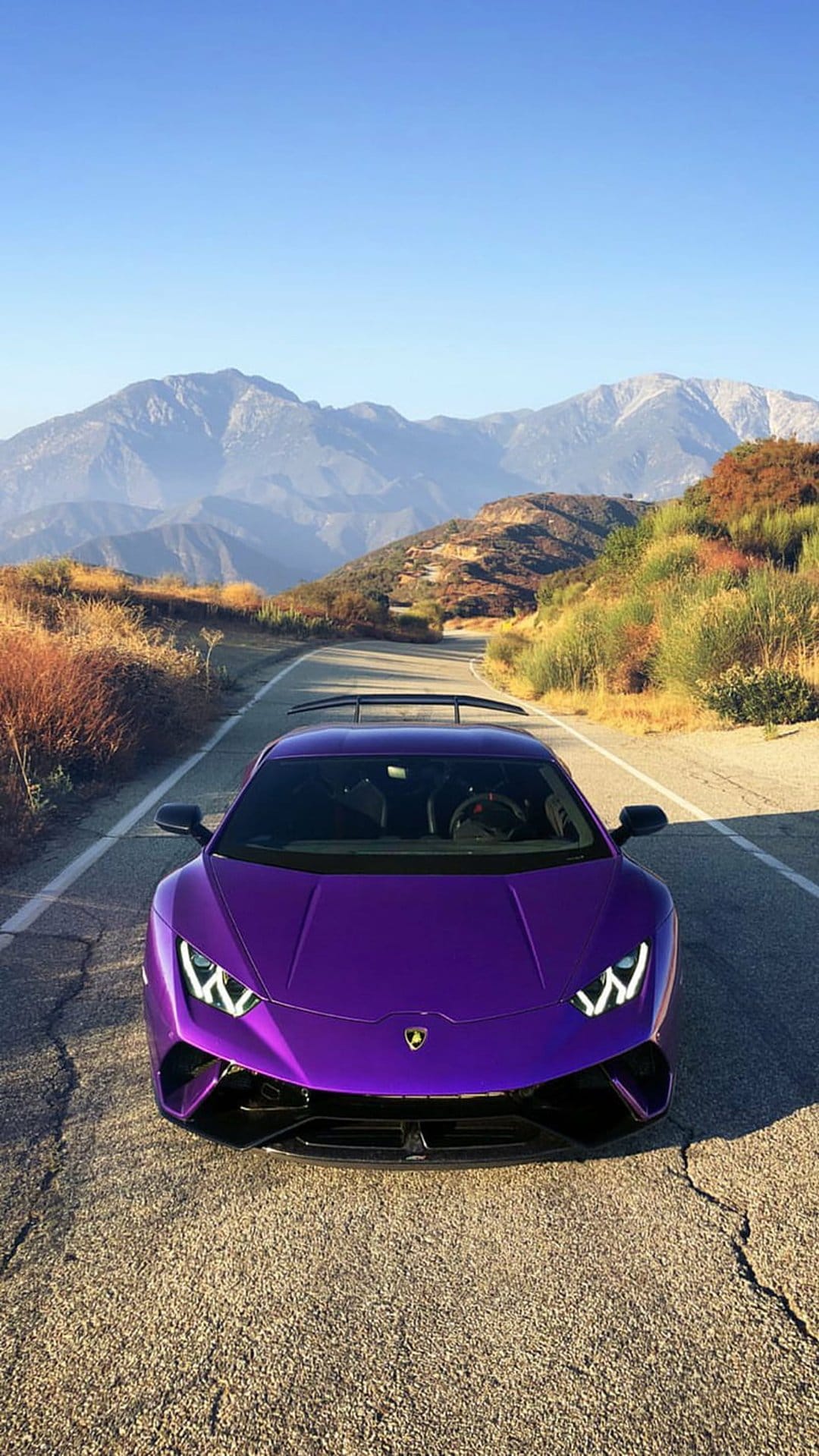 Purple Lamborghini supercar parked on a winding asphalt road with green hills and mountains in the background.