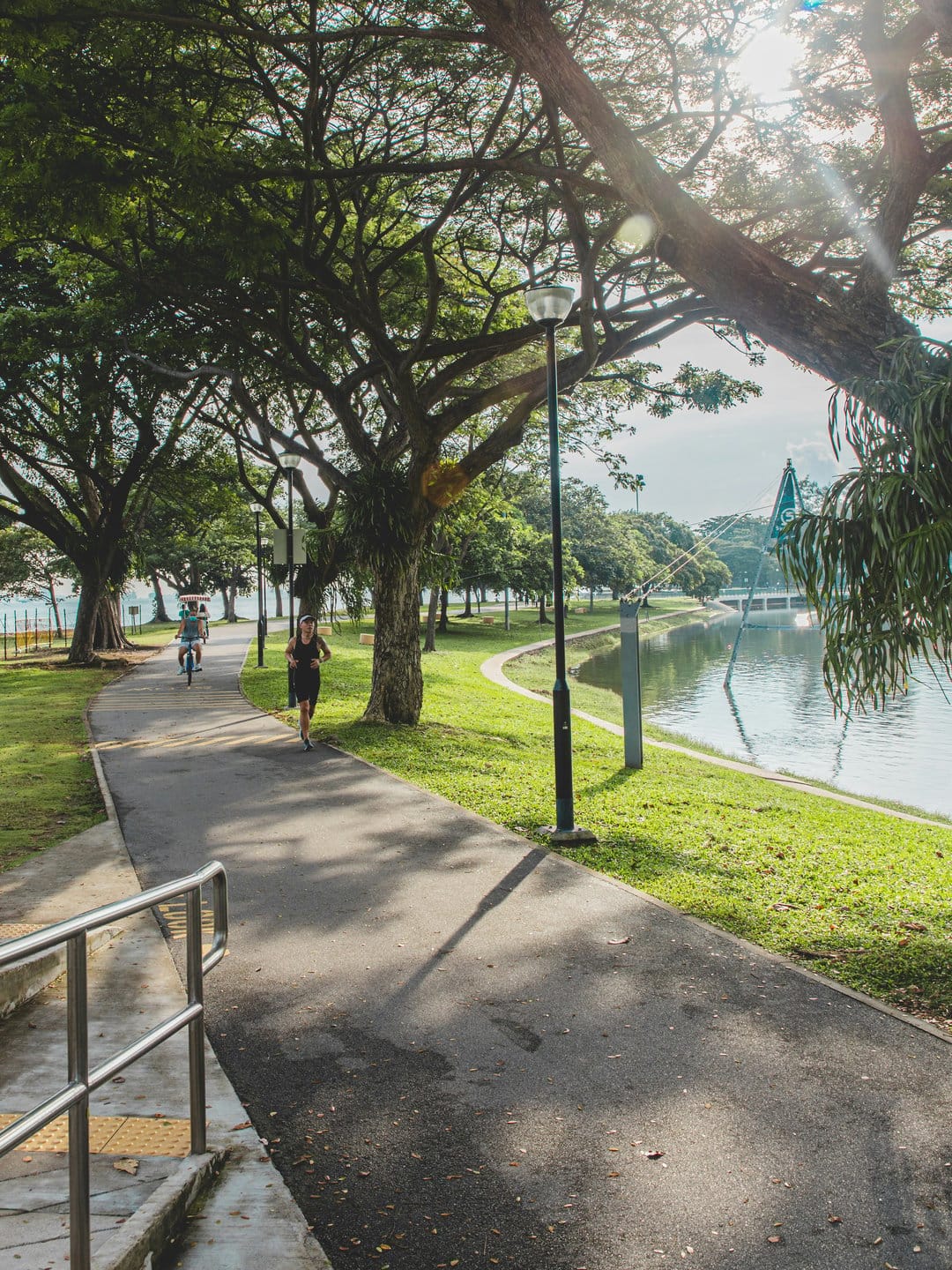 Woman in athletic gear jogging on a paved park path beside a calm lake with sunlight filtering through large trees.