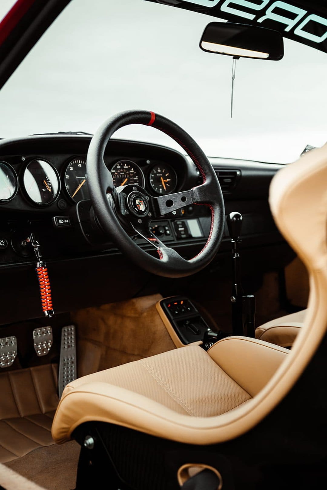 Porsche sports steering wheel with red stitching and tan leather racing seats inside a vintage car cabin.