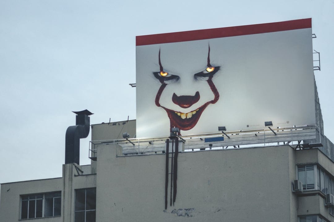 Pennywise the clown with glowing yellow eyes on a rooftop billboard against a cloudy, dark urban skyline.