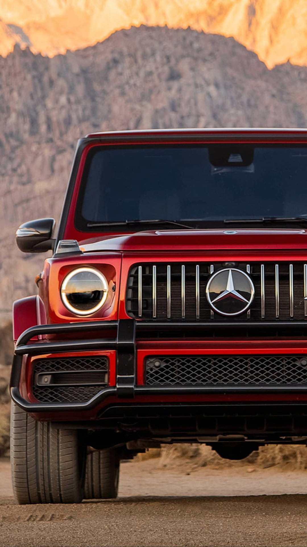 Red Mercedes G-Wagon SUV with circular headlights parked in a rocky desert landscape under a warm sunset sky.
