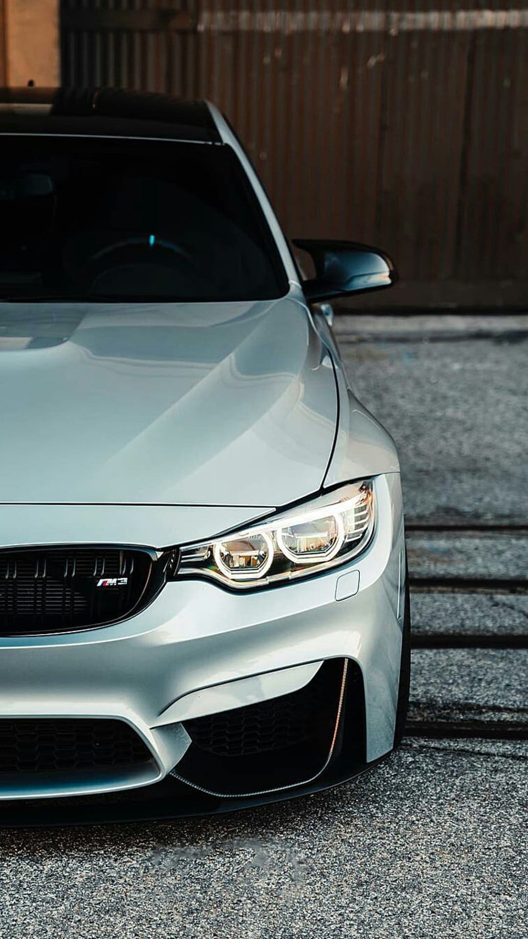 Silver BMW M4 front bumper featuring sharp LED headlights and a black M grille against a rustic wooden wall.