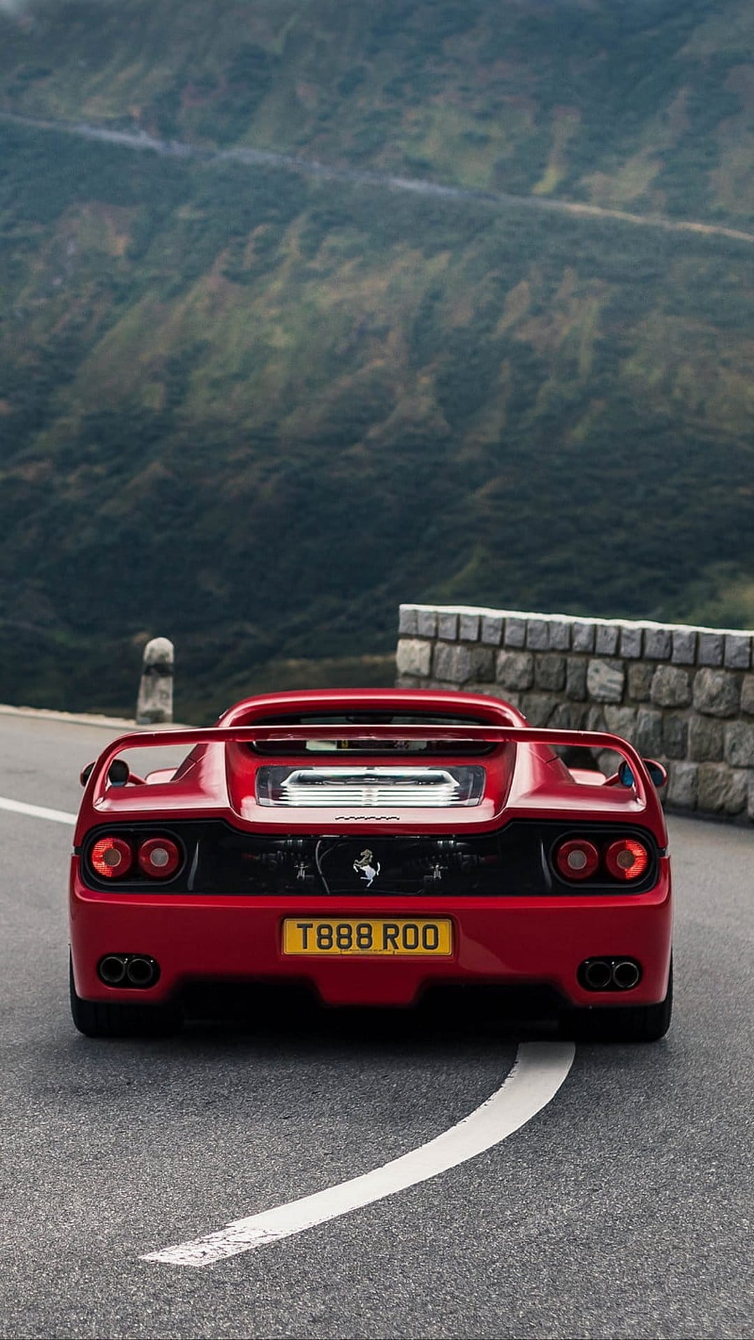Red Ferrari F50 rear view parked on a mountain road with green hills and a stone wall in the background.