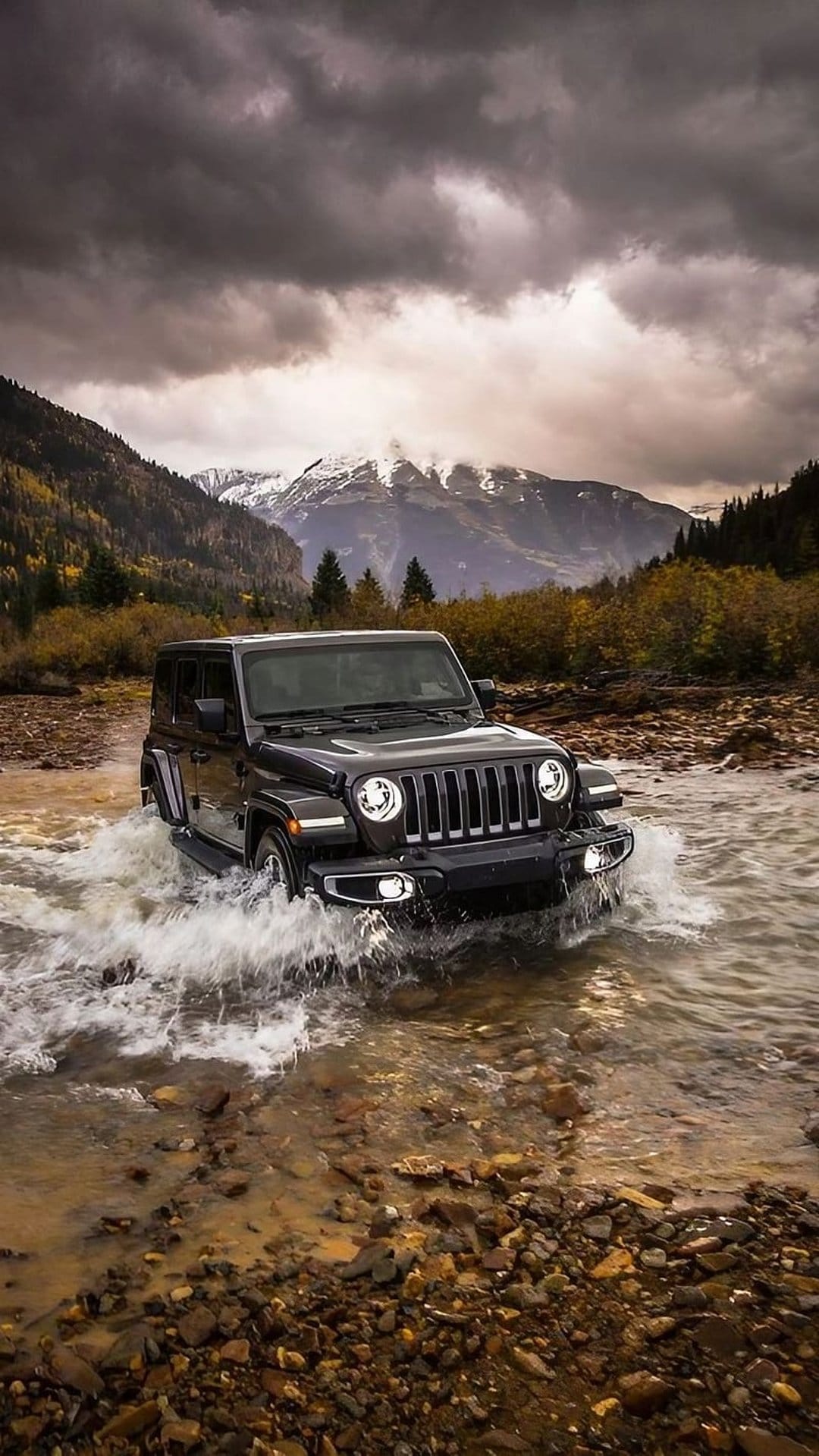 Jeep Wrangler splashing through a rocky mountain river under heavy storm clouds and distant snowy peaks.
