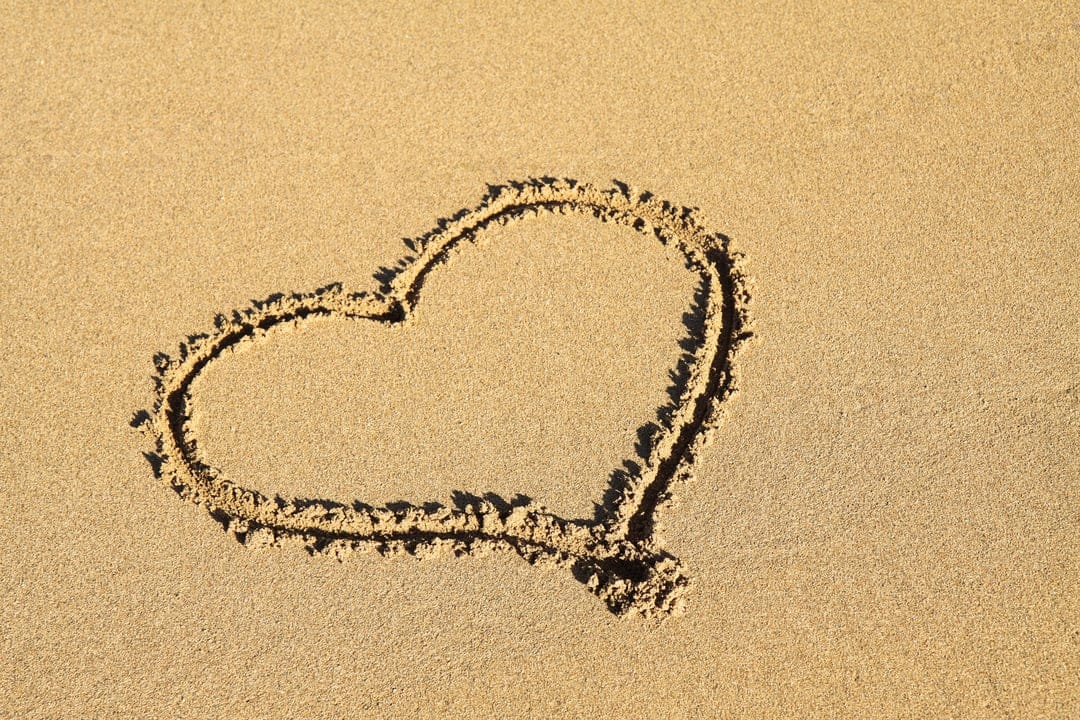 Heart shape hand-drawn in textured beach sand illuminated by warm golden sunlight during a coastal sunset.
