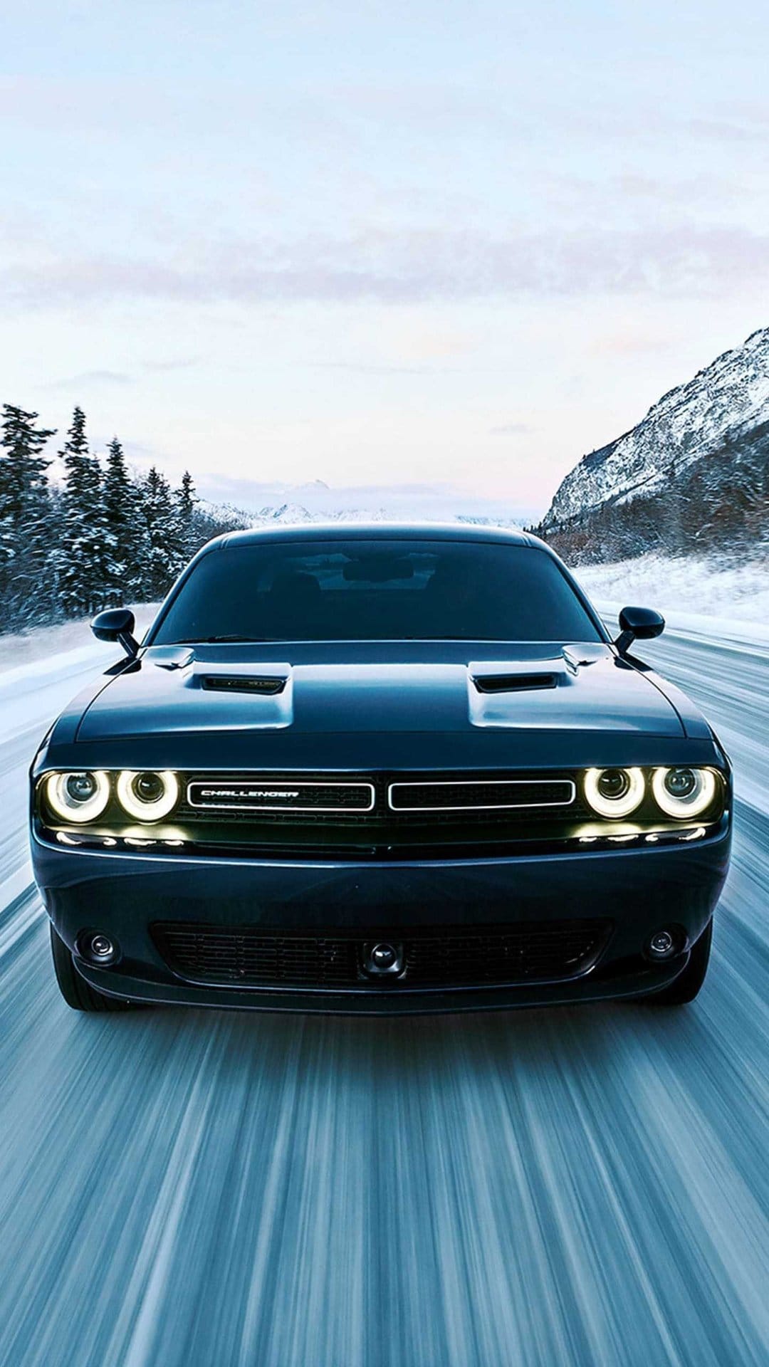 Black Dodge Challenger with glowing halo headlights driving on a snowy mountain road with pine trees.