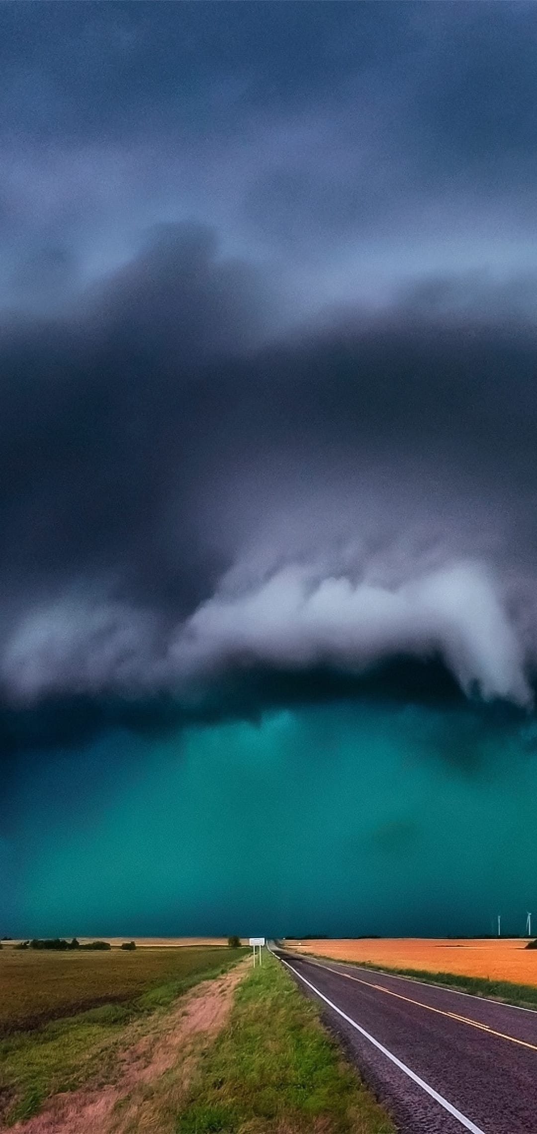 Dark storm clouds hover over a straight asphalt road and a teal horizon in a dramatic rural landscape scene.