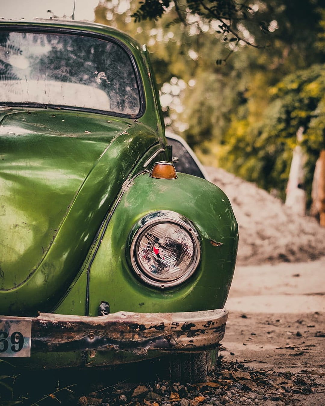 Green classic car headlight and chrome bumper with visible dust and scratches against a blurred forest backdrop.