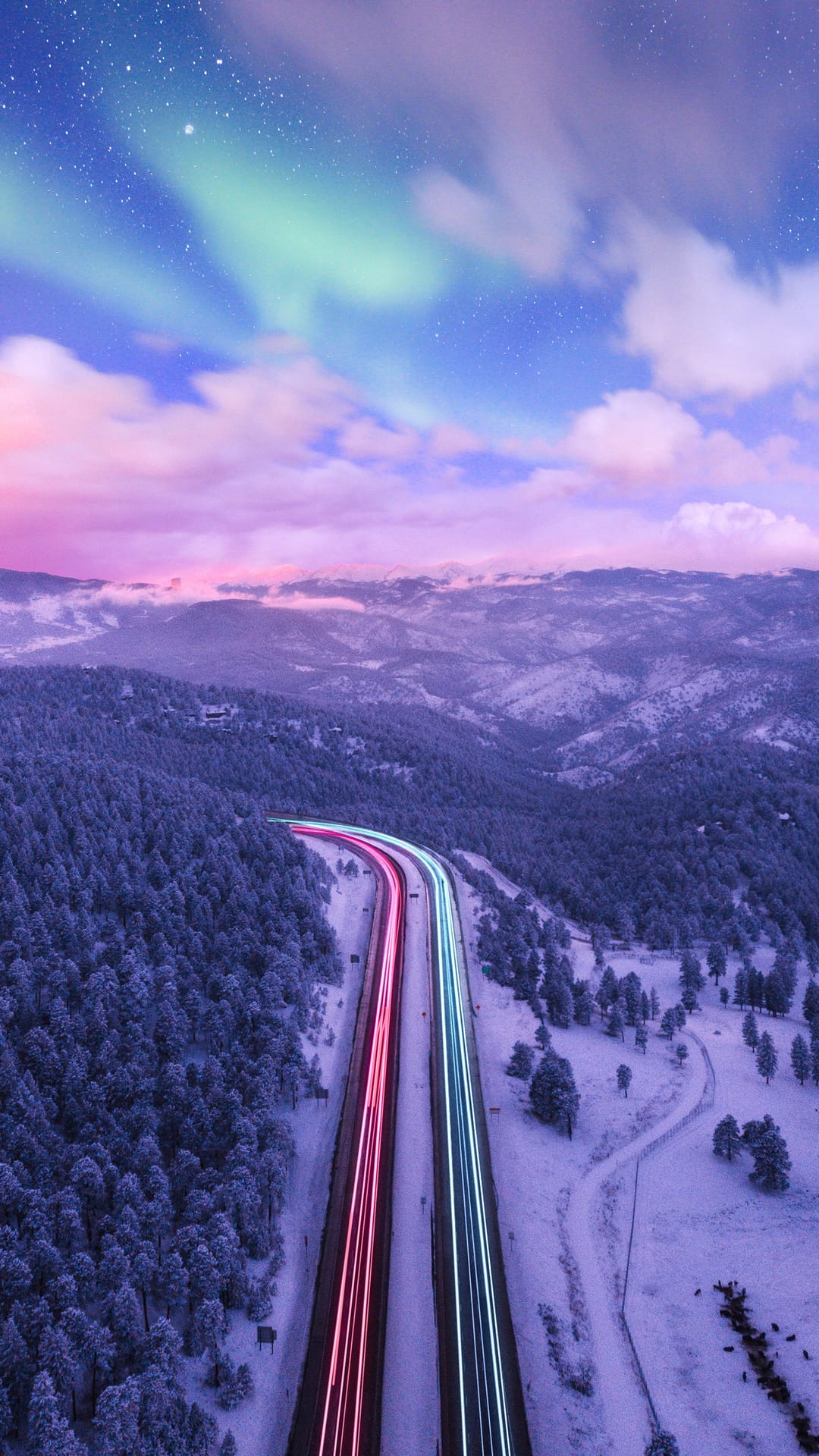Winding snowy road with red and blue light trails through a pine forest under a pink and green aurora sky.