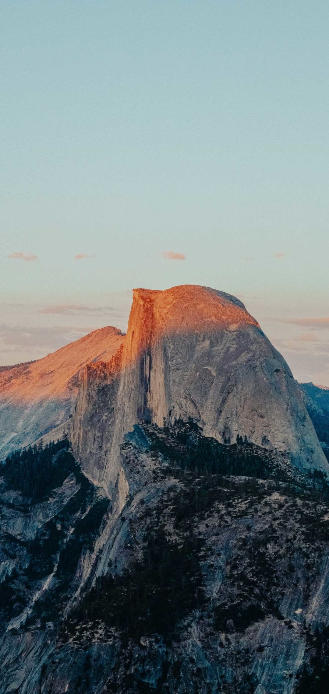 Half Dome's granite face glows orange at dusk above forested mountains under a soft pink and purple pastel sky.
