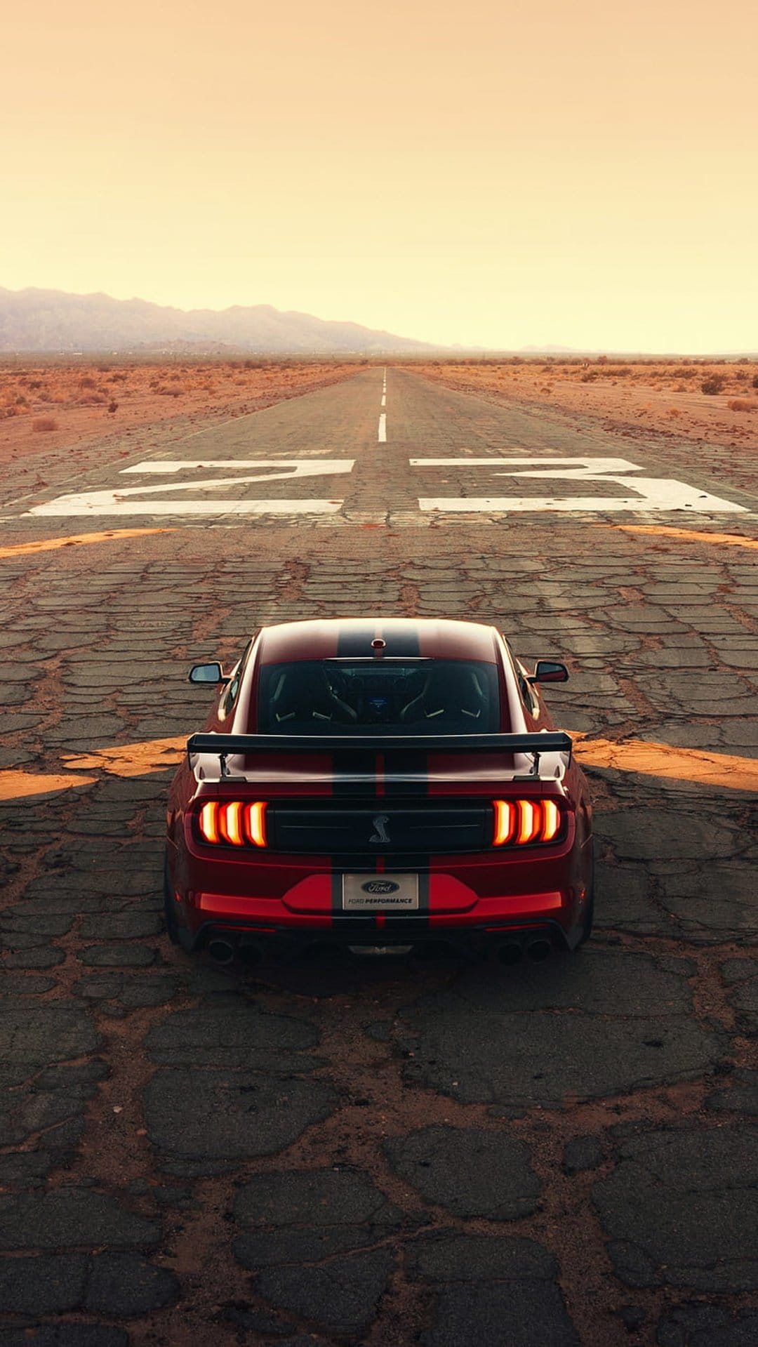 Red Shelby GT500 muscle car parked on a cracked desert runway under an orange sunset with glowing brake lights.