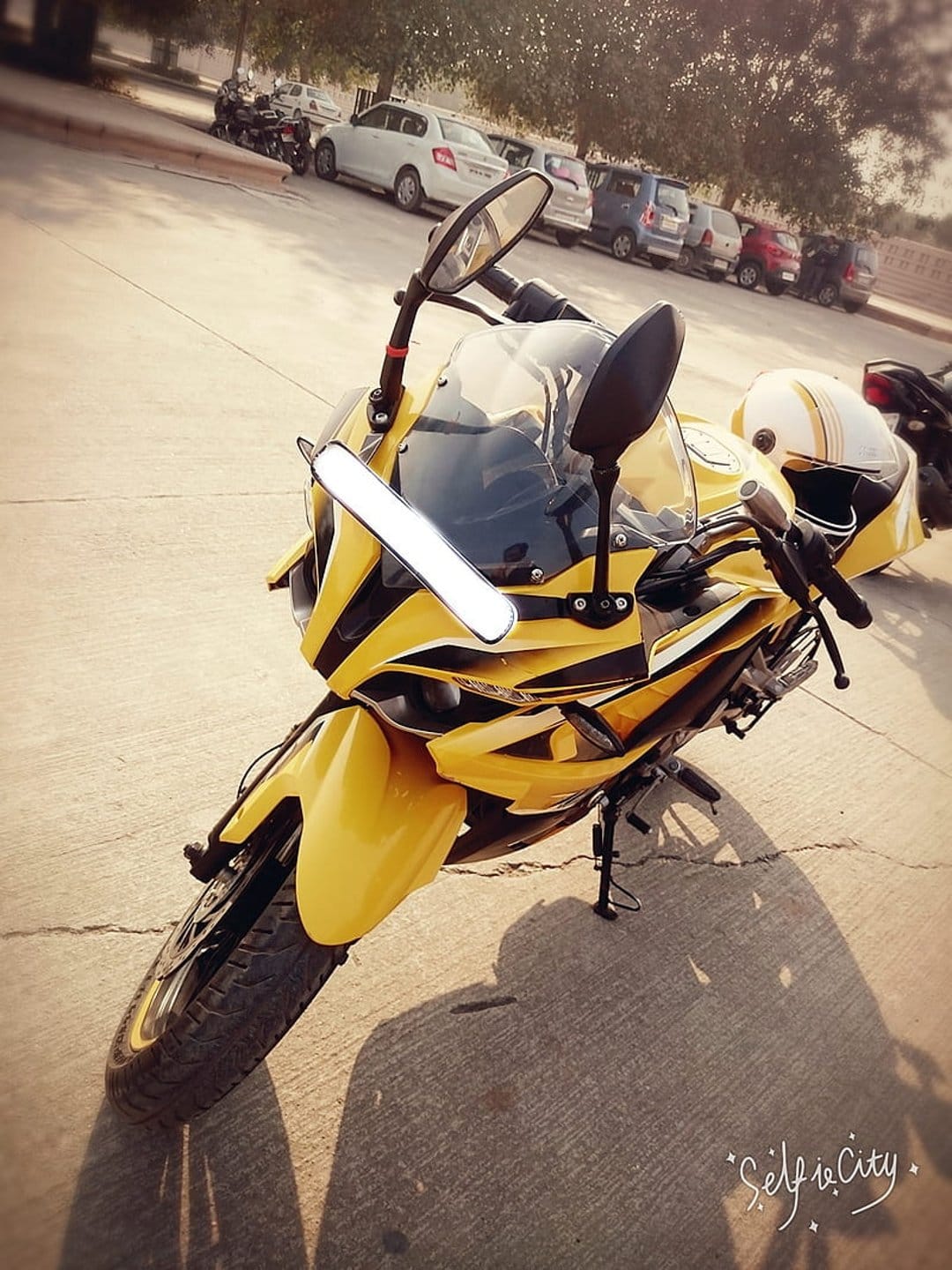 Yellow sport motorcycle with a helmet on the seat parked on sunny asphalt with trees and cars in the background.