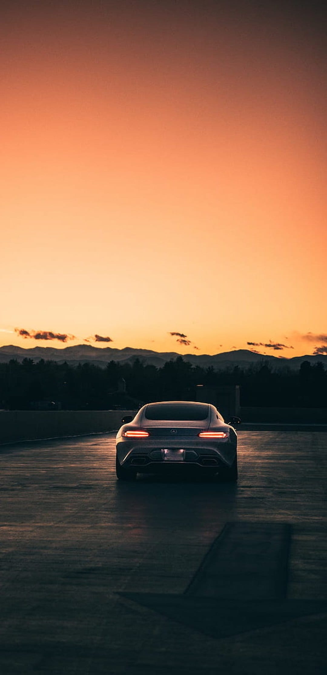 Luxury sports car with red taillights parked on asphalt during a mountain sunset with an orange and purple sky.
