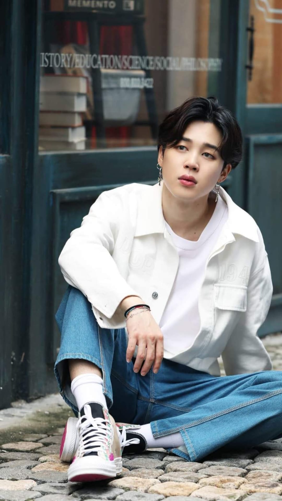 Jimin of BTS sits on cobblestone wearing a white jacket, blue jeans, and sneakers in a vertical portrait.
