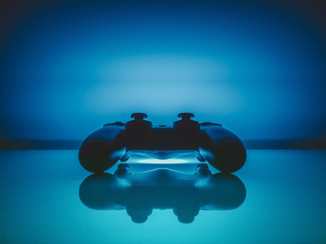 Black gaming controller with glowing blue internal lights sits on a glossy dark surface with a clear reflection.