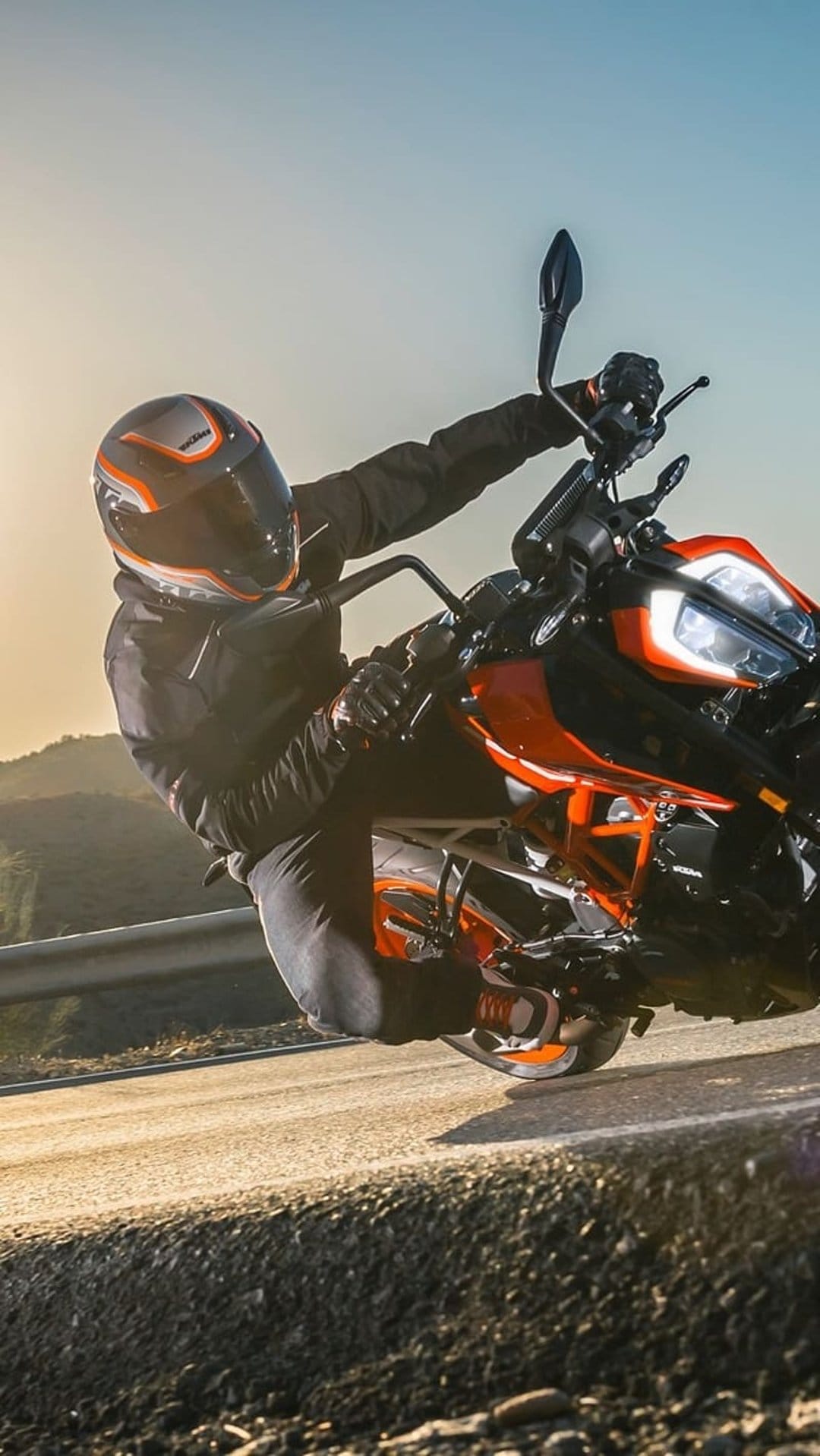 Motorcyclist in black gear leans an orange sportbike into a sharp road turn against a sunset mountain backdrop.